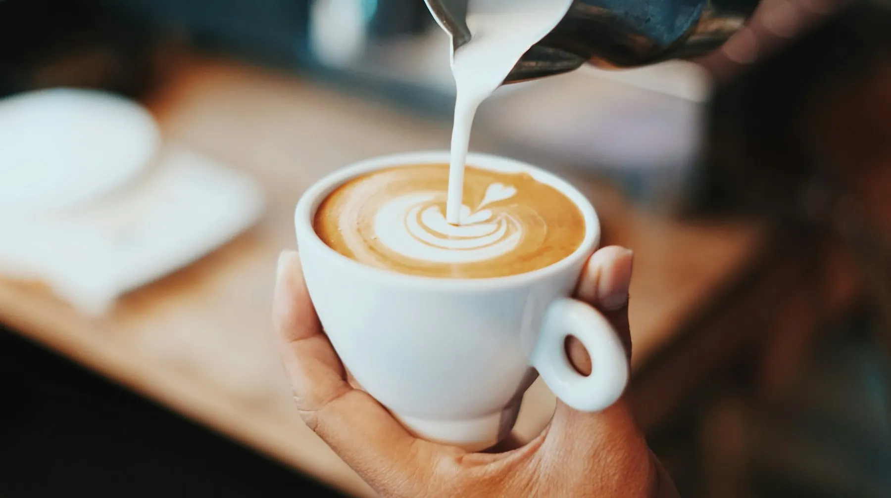Barista toarnă lapte texturat într-o ceașcă de cappuccino ținută în mână